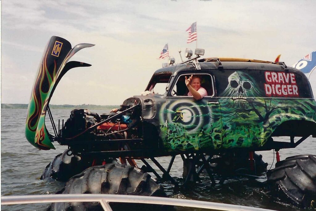 A man waves from the drivers seat of a black old-time vehichle with huge tires in water, hand painted with a green haunted forest.