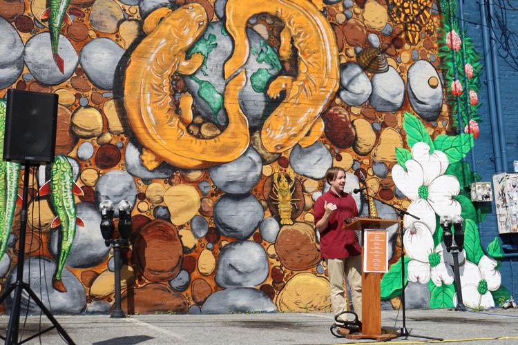 Boone Town Council Member Dalton George, wearing a maroon shirt and khaki pants, speaks at an outdoor podium with a microphone in front of a large, colorful mural depicting two orange Hellbender salamanders intertwined among rocks and aquatic plants. The mural, painted on a brick wall, also features fish, crayfish, and blooming white flowers with green leaves. A black speaker on a stand and two parking meters are visible in the foreground. The event takes place in daylight, with bright sunlight casting shadows.